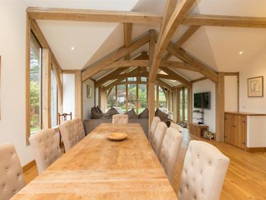 A dining room with a long wooden table and beige upholstered chairs near wooden beams and large windows at Old Droomer in Windermere