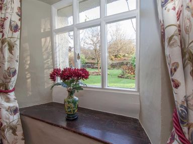 A window with floral curtains and a vase of red flowers on the sill overlooking a garden at Old Droomer in Windermere