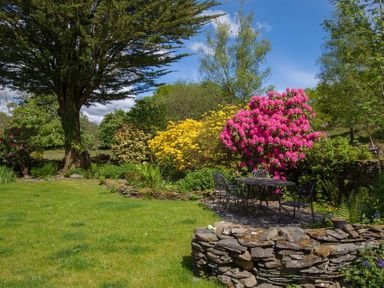 A garden with trees colorful flowering bushes stone wall and outdoor metal table and chairs at Old Droomer in Windermere