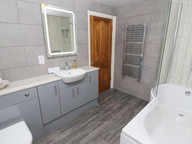 A bathroom with a wooden door gray cabinets a white sink a lit mirror a chrome towel rack and a white bathtub at Kirkstones in Keswick