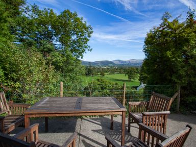 An outdoor patio with a wooden table and chairs overlooking a green field with trees and hills in the distance at Kirkstones in Keswick