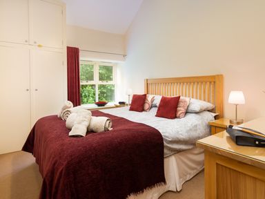 A bedroom with a bed covered in white and burgundy bedding with rolled towels on top near a window with red curtains at Tethera in Keswick