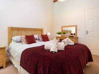 A bedroom with a wooden bed frame burgundy bedding and rolled towels on the bed a wooden dresser with a mirror and a plant and a closed door at Tethera in Keswick