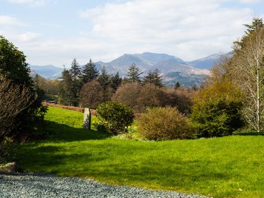 A garden with grass shrubs and trees overlooking mountains under a partly cloudy sky at Tethera in Keswick