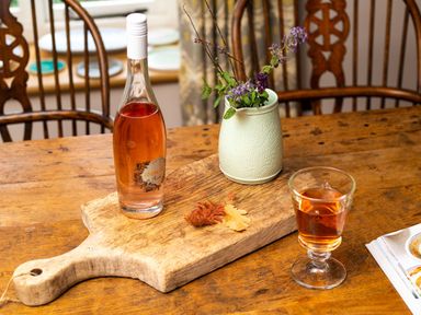 A wooden dining table with a bottle of rose wine on a wooden cutting board next to a ceramic vase with flowers and a glass of rose wine at High Cunsey Farm in Far Sawrey