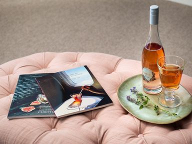 A pink tufted cushion with two books stacked and a plate holding a bottle of rose wine a glass of wine and some sprigs of herbs at High Cunsey Farm in Far Sawrey