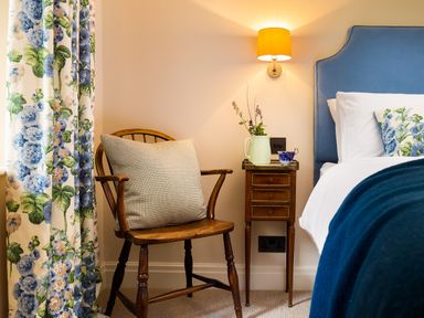 A bedroom corner with a wooden chair with a cushion next to a bedside table with a lamp a jug with flowers and a teacup at High Cunsey Farm in Far Sawrey