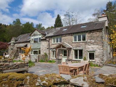 A stone house with multiple windows and a wooden outdoor table with chairs on a stone patio at High Cunsey Farm in Far Sawrey