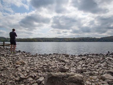 A man standing on a rocky shore looking at a lake with hills and trees in the background at High Cunsey Farm in Far Sawrey
