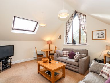 A living room with a sofa, coffee table, TV, small dining table with chairs, floor lamp, and triangular window with curtains at Squirrel Bank in Ambleside