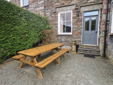 An outdoor area with a wooden picnic table and benches beside a stone building with a grey door and window surrounded by green bushes at Fairy Glen in Grasmere near Ambleside