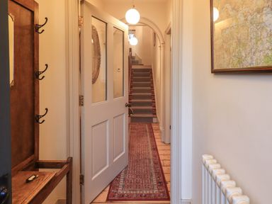 A hallway with a white door open showing a carpet runner on wooden floor leading to stairs with a blue runner and round ceiling lights at Fairy Glen in Grasmere near Ambleside