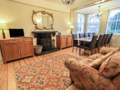 A living room with a patterned rug a floral sofa a wooden dining table with chairs two wooden cabinets lamps a fireplace with a mirror above it and large windows at Fairy Glen in Grasmere near Ambleside