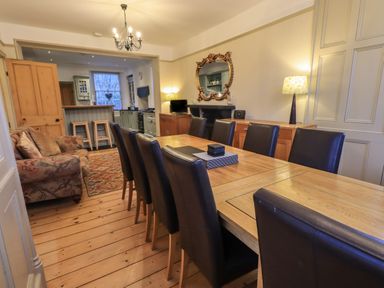 A dining room with a long wooden table and black chairs next to a sitting area and open kitchen at Fairy Glen in Grasmere near Ambleside