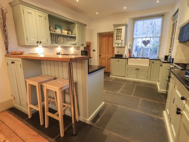 A kitchen with light green cabinets and black countertops two wooden stools at a wooden breakfast bar at Fairy Glen in Grasmere near Ambleside