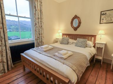 A bedroom with a wooden bed frame two bedside tables with lamps a window with floral curtains and a mirror above the bed at Fairy Glen in Grasmere near Ambleside