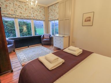A bedroom with a bed with folded towels a rug two chairs a chest of drawers and floral window blinds at Fairy Glen in Grasmere near Ambleside