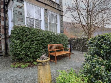An outdoor area with a wooden bench near a stone house with large windows and green bushes at Fairy Glen in Grasmere near Ambleside