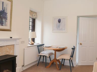 A small dining area with a wooden table, two blue chairs with cushions, a lamp on a windowsill, and framed artwork on the walls at Larch Cottage in Hawkshead