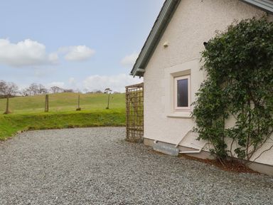 The exterior side of a house with a small window climbing plants a trellis and a gravel area with a grassy hill in the background at Larch Cottage in Hawkshead