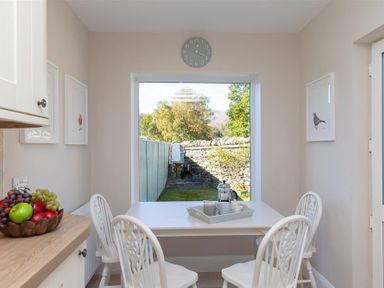 A dining area with a white table and four chairs next to a large window showing a garden at Crozier Cottage in Grasmere