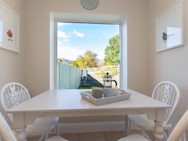 A dining area with a white table and chairs next to a large window with a view of a garden at Crozier Cottage in Grasmere