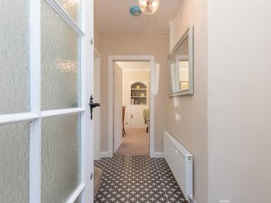 A hallway with patterned floor tiles a radiator and a mirror leading to a room with shelves and chairs at Crozier Cottage in Grasmere