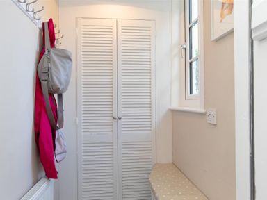 A small hallway with a coat rack, a gray bag, a red jacket, a window, a radiator, and white louvered doors at Crozier Cottage in Grasmere