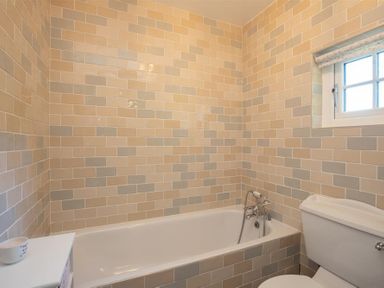A bathroom with a bathtub toilet window and beige and gray tiled walls at Crozier Cottage in Grasmere