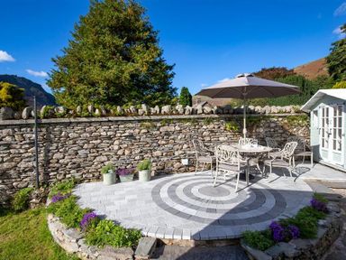 An outdoor patio with a circular patterned floor table and chairs under an umbrella near a stone wall at Crozier Cottage in Grasmere