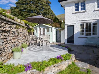 An outdoor patio area with a round table and chairs under an umbrella next to a stone wall and a small shed at Crozier Cottage in Grasmere