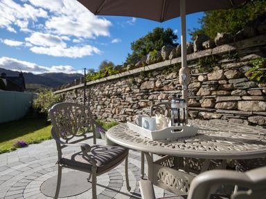 A patio with metal chairs and a table with a tray and coffee set under an umbrella by a stone wall at Crozier Cottage in Grasmere