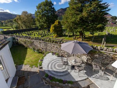 An outdoor patio with chairs a table and umbrella overlooking a graveyard surrounded by trees at Crozier Cottage in Grasmere