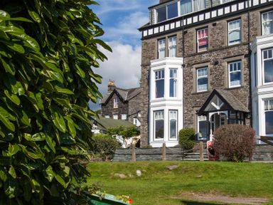 A stone house with white bay windows and a porch surrounded by grass and bushes at Waterside in Ambleside