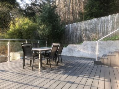 An outdoor deck with a table and six chairs next to stairs and a wooden fence with trees behind at Dancing Beck Cottage in Millbeck