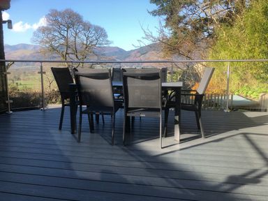 A balcony with a black dining table and six chairs with glass railing and mountains in the background at Dancing Beck Cottage in Millbeck