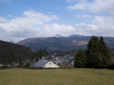 A landscape with a grassy field in the foreground houses and trees in the middle and mountains with snow patches in the background at Bandol in Braithwaite