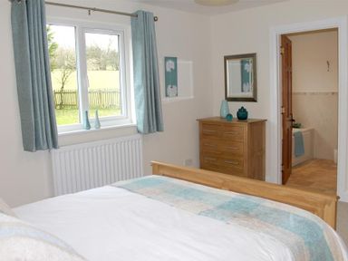 A bedroom with a wooden bed frame a window with curtains a wooden dresser and an open door showing a bathroom at Bandol in Braithwaite