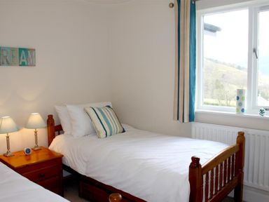 A bedroom with two single wooden beds one with a striped pillow a wooden bedside table with two lamps a clock and a window with curtains at Bandol in Braithwaite