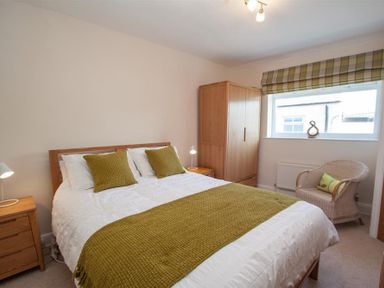A bedroom with a double bed green cushions and throw two bedside tables wooden wardrobe wicker chair and window at Gable End in Braithwaite