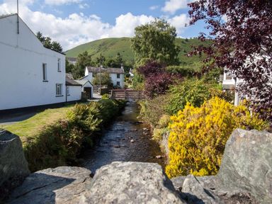 A stream flowing between bushes and houses with a hill and partly cloudy sky in the background at Gable End in Braithwaite