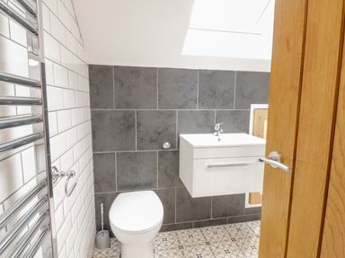 A bathroom with a white toilet a white sink with a cabinet patterned floor tiles grey wall tiles and a wooden door at Meadow View in Tyn-y-Gongl near Benllech