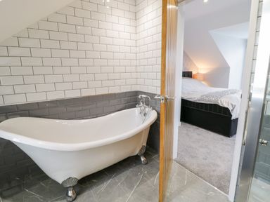 A bathroom with a clawfoot bathtub and tiled walls adjoining a bedroom with a bed and a bedside lamp at Meadow View in Tyn-y-Gongl near Benllech