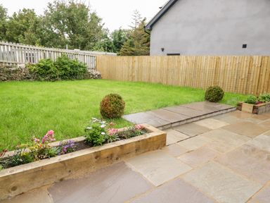 A backyard with a green lawn wooden flower beds and a paved patio at Meadow View in Tyn-y-Gongl near Benllech