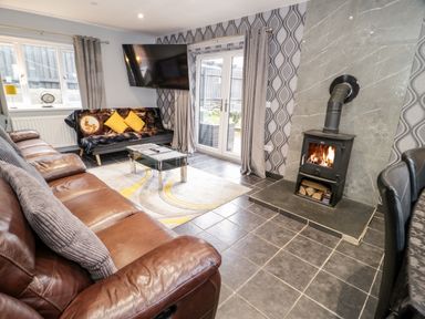 A living room with a brown leather sofa a black futon with yellow cushions a glass coffee table a wood burning stove and a wall mounted television at Meadow View in Tyn-y-Gongl near Benllech