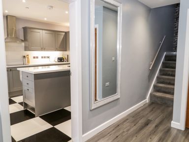 A hallway with a large wall mirror and a staircase next to a kitchen with an island and checkered floor at Meadow View in Tyn-y-Gongl near Benllech