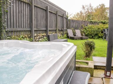A bubbling hot tub with wicker lounge chairs and a bench on grass surrounded by a wooden fence