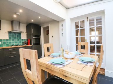 A kitchen with a wooden dining table set with plates glasses and cutlery and a modern cooking area with teal tiles at South Harbour Cottage in Weymouth