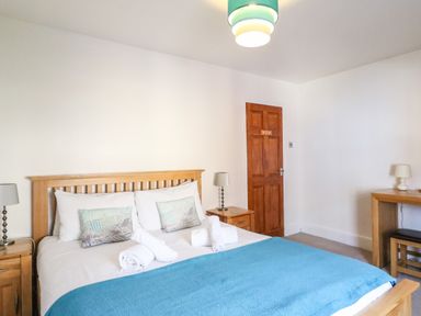 A bedroom with a double bed with blue blanket and rolled towels bedside tables lamps and a wooden desk with a stool at South Harbour Cottage in Weymouth
