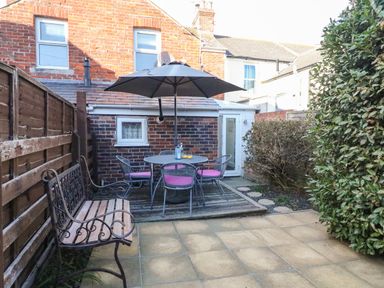A backyard patio with a metal bench a round table with pink cushioned chairs and an umbrella at South Harbour Cottage in Weymouth